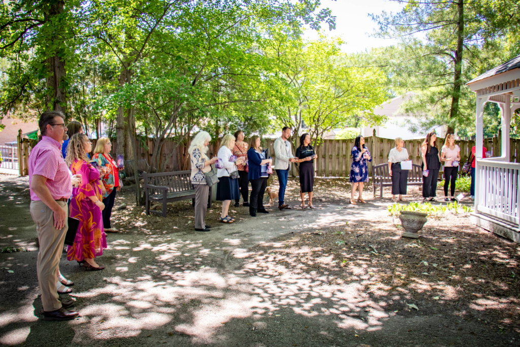 Group of people standing outdoors in a shaded garden area near benches and a white gazebo.