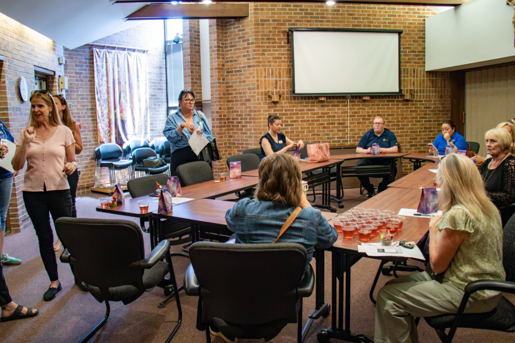 Group of people seated around tables with drinks in a brick-walled meeting room with a projector screen.