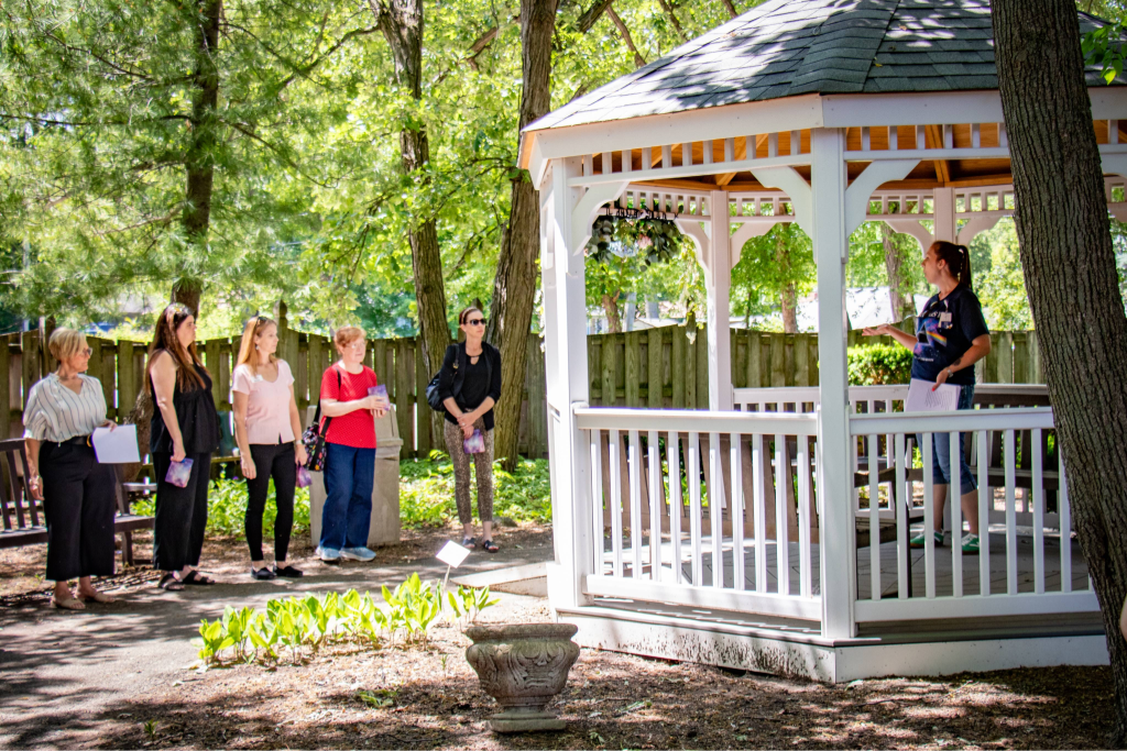 Woman standing in a white gazebo speaking to five women lined up outside in a shaded garden area.