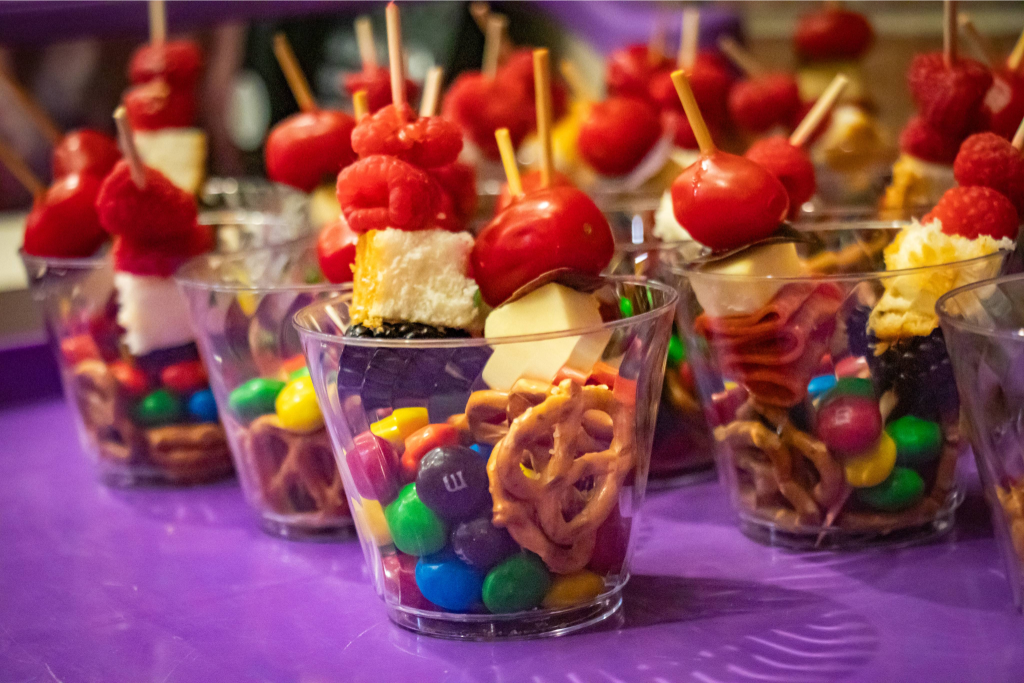 Clear cups filled with colorful candies, pretzels, and skewers topped with raspberry and cherry.