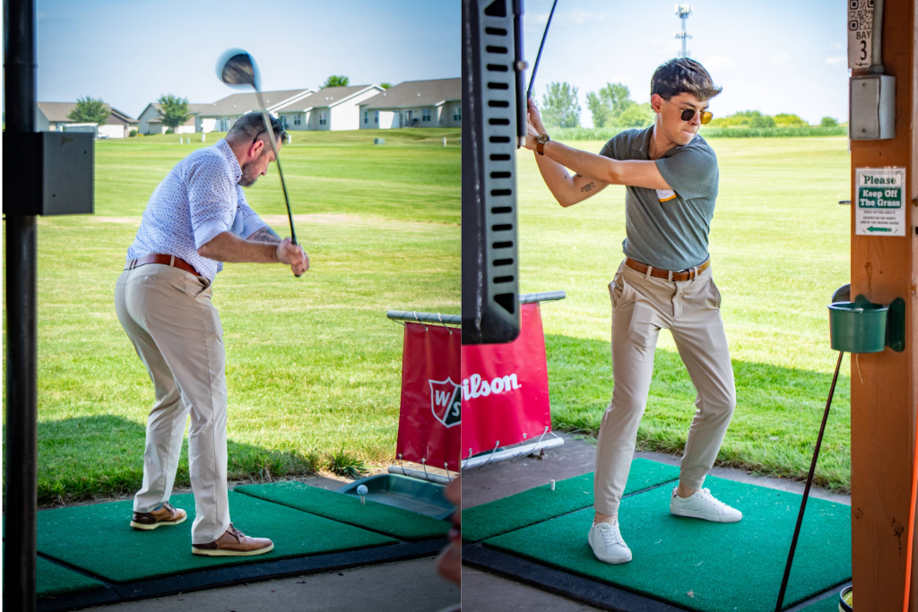Two men practice their golf swings at a driving range on a sunny day with green grass and houses behind.