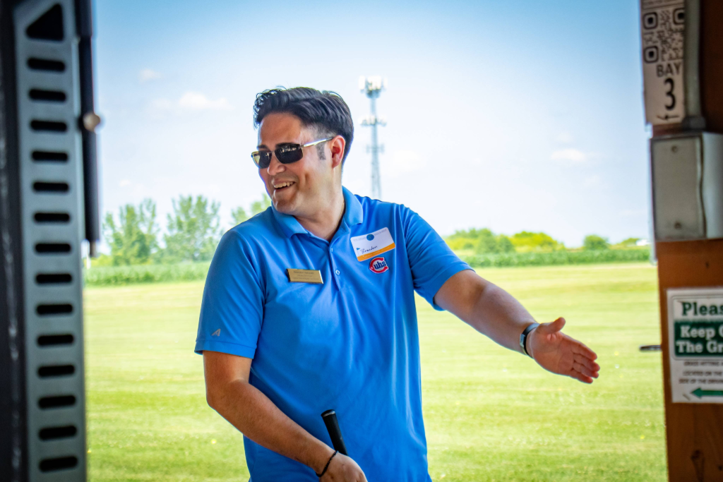 Man in blue polo and sunglasses smiling and gesturing with one hand outside on a sunny day.