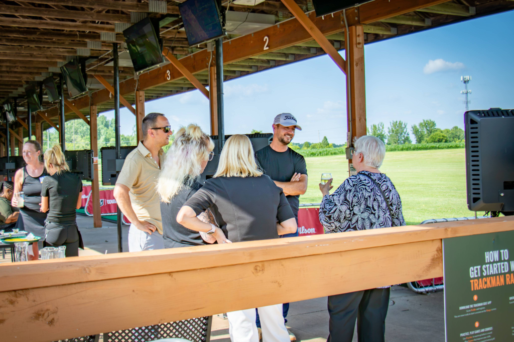 Group of people socializing under a wooden roof at a golf event with green field in the background.