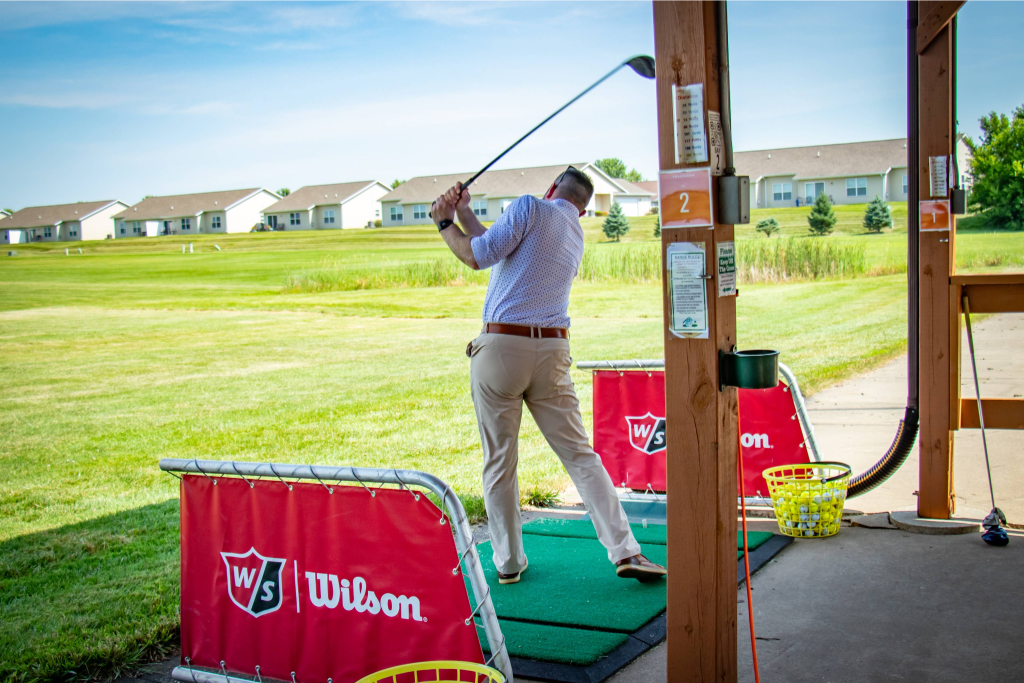 Man swinging golf club at driving range with Wilson-branded barriers and a basket of golf balls nearby.