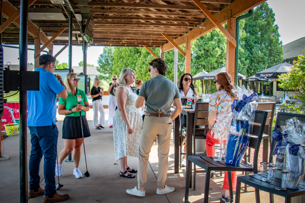 Group of people socializing under wooden roof at an outdoor golf event with gift baskets nearby.