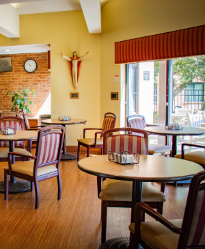 Empty dining area with round tables, striped chairs, a crucifix on the wall, and large windows with red valance.