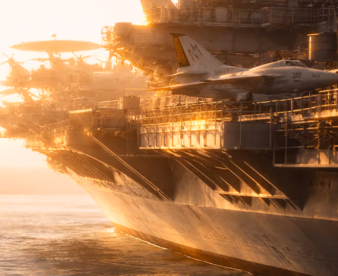 Side view of a navy aircraft carrier at sunset with a fighter jet on the deck.