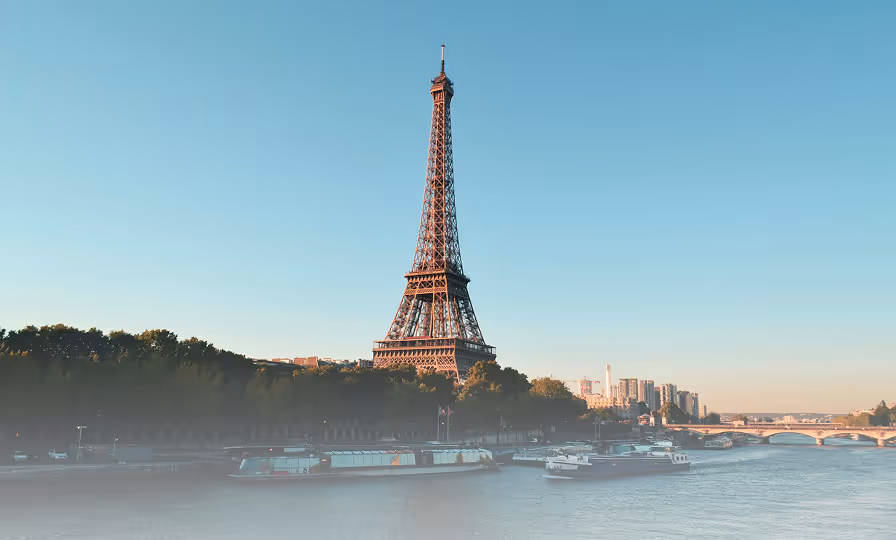 Eiffel Tower standing tall near the Seine River with boats and a tree-lined riverbank under a clear blue sky.