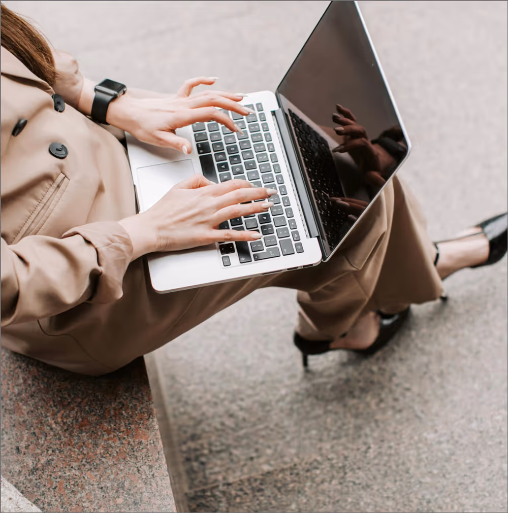 A woman typing on a laptop while sitting on steps in neutral toned outfit