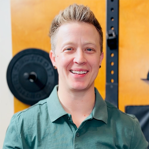 Smiling person with short, light hair wearing a green collared shirt in a gym setting with a weight rack in the background.