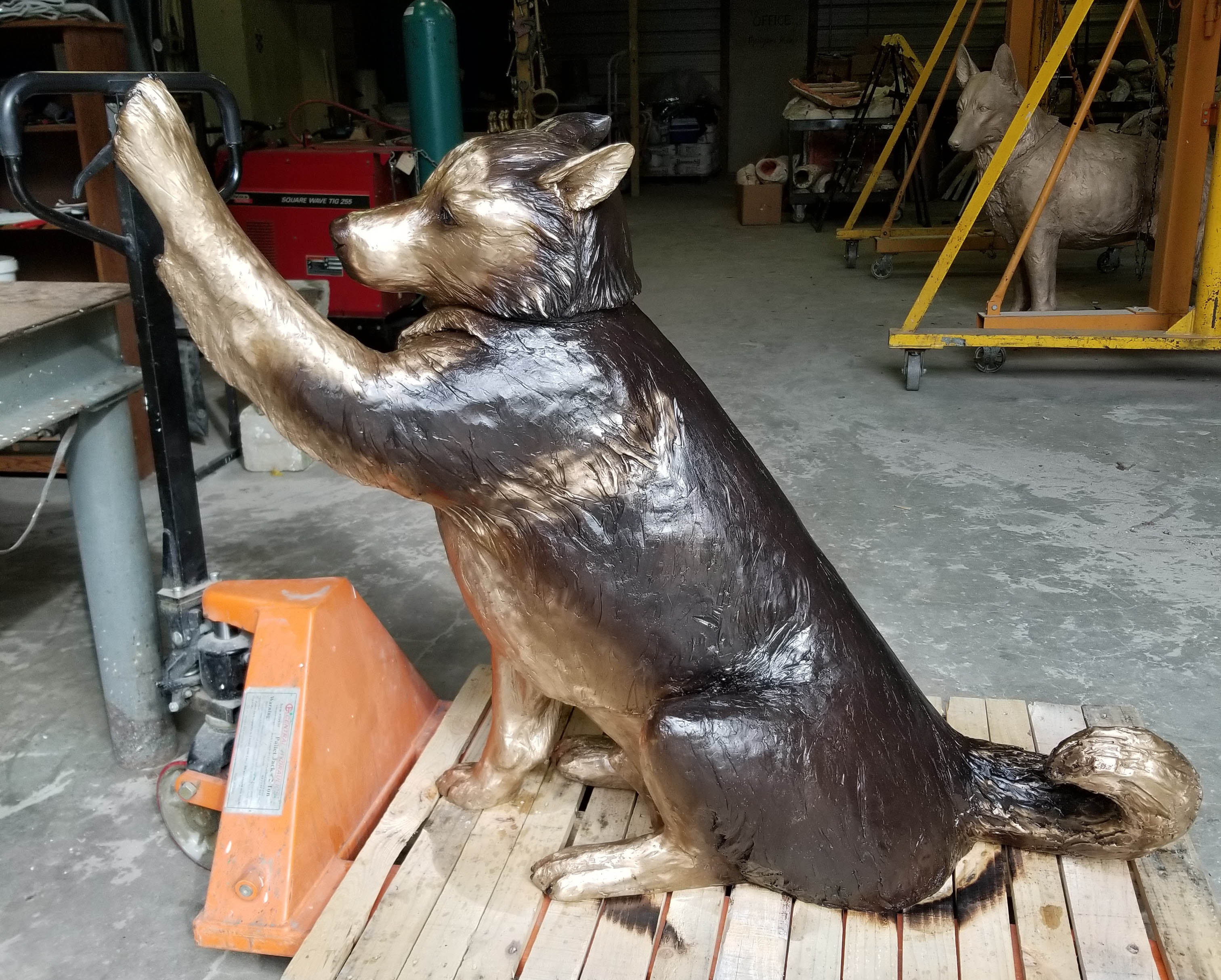Bronze sculpture of a seated dog with raised front paws on a wooden pallet in a workshop.