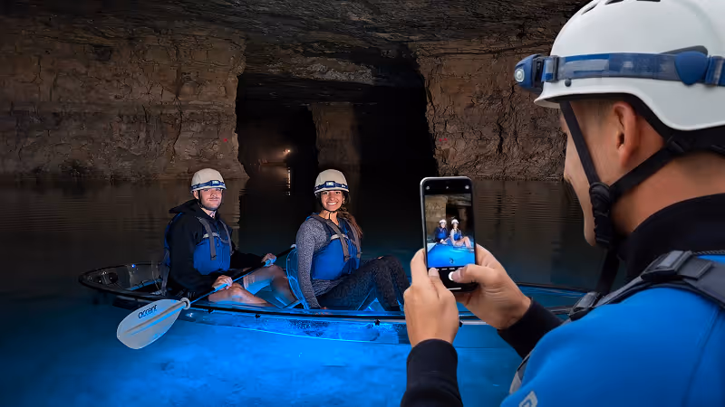 Tour guide taking a photo of guests in a clear-bottom kayak in a cave with blue glowing underneath in the water.