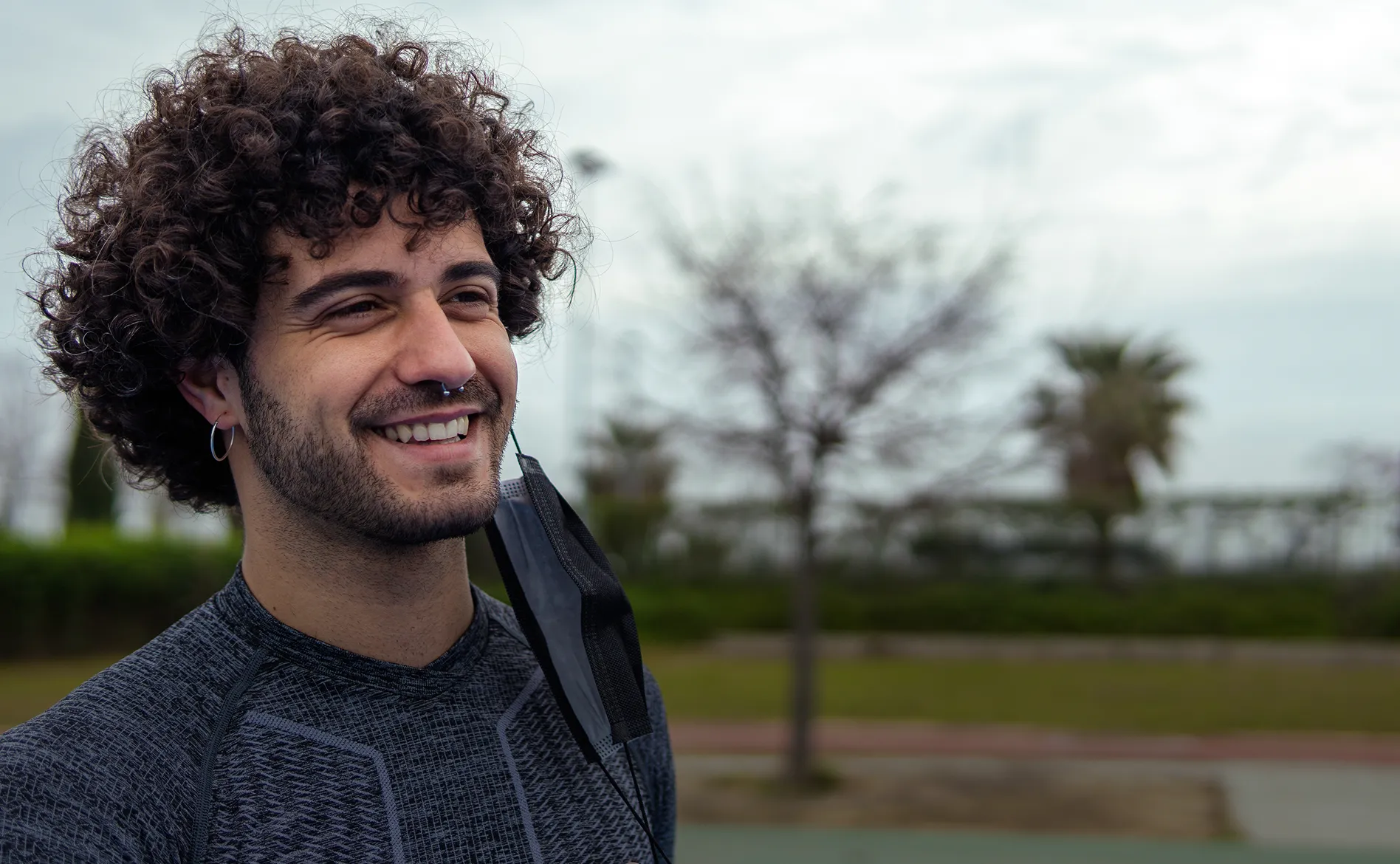Smiling young man with curly hair and septum piercing wearing a dark patterned shirt and a black face mask hanging from his ear, outdoors near trees.