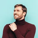 Man with short brown hair and beard smiling, wearing a burgundy turtleneck sweater against a light blue background.