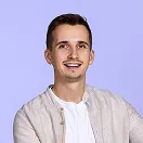 Smiling young man with short brown hair and mustache, wearing a beige jacket over a white shirt, against a light purple background.