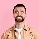 Smiling man with short dark hair, beard, and tan shirt against a pink background.