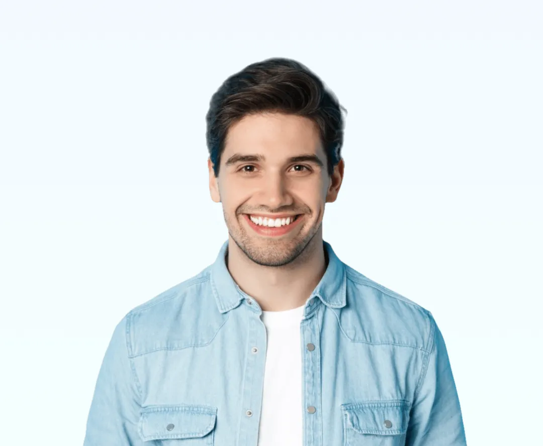 Smiling young man with dark hair wearing a light denim shirt over a white t-shirt on a light blue background.