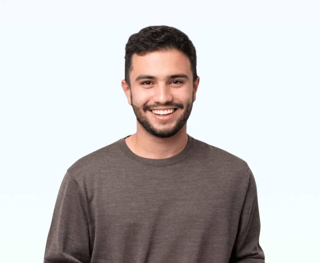 Smiling young man with short dark hair and beard wearing a brown sweater against a light background.