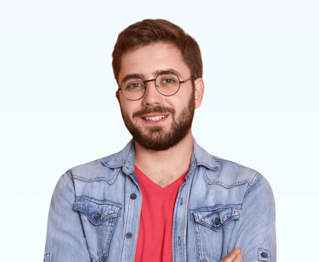 Young man with beard and glasses wearing a red shirt and denim jacket, smiling with arms crossed.