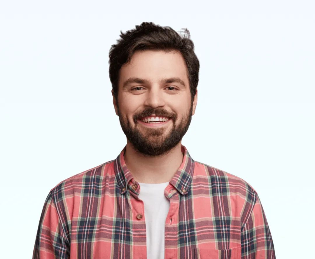 Smiling man with dark hair and beard wearing a red and blue plaid shirt over a white t-shirt.