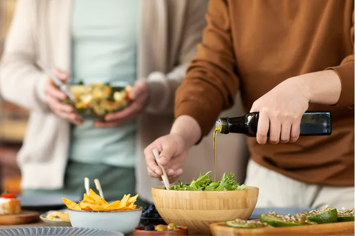 Person pouring olive oil from a dark bottle over a green salad in a wooden bowl.
