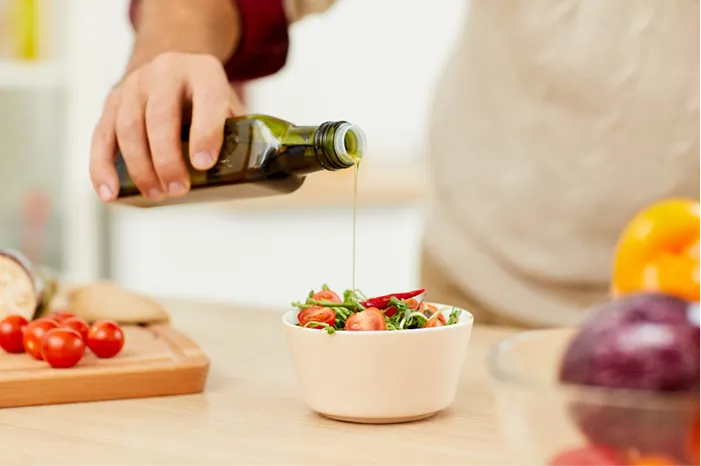 Hand pouring olive oil from a green bottle onto a small bowl of salad.