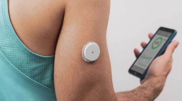 Close-up of a round white CGM sensor on a person's upper arm, with a smartphone displaying a glucose monitoring app in the background.
