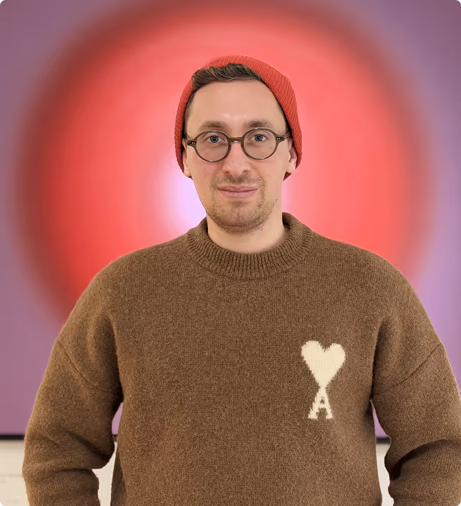 Man wearing round glasses, a red beanie, and a brown sweater with a white heart and letter A design, standing in front of a blurred red and purple background.