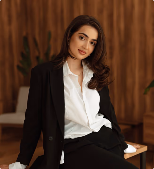 Woman with long brown hair wearing a white shirt and black blazer sitting on a wooden bench against a brown wooden wall background.