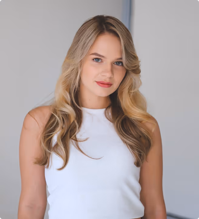 Blonde woman with loose curls wearing a white sleeveless top looking at the camera against a neutral background.