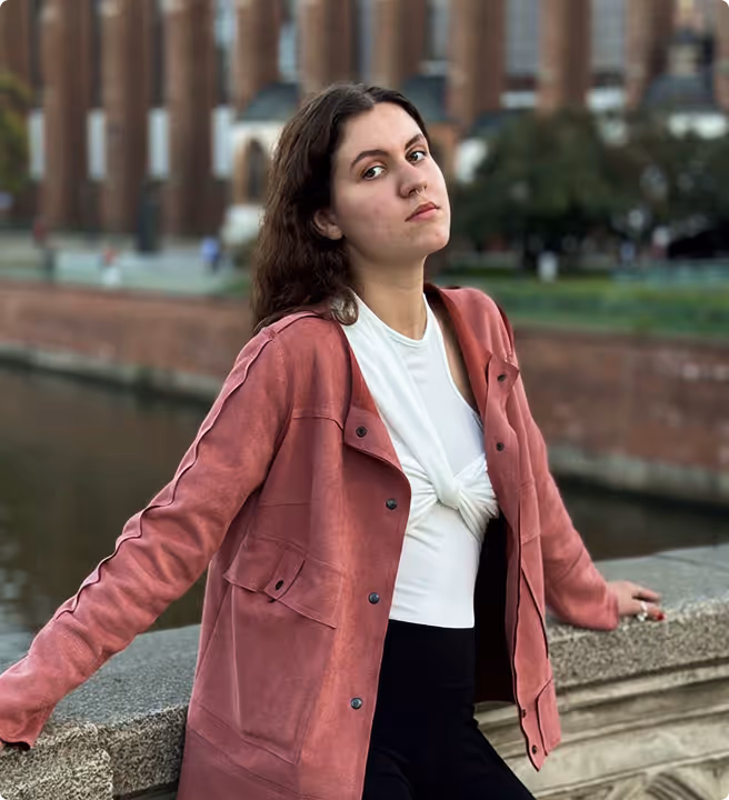Young woman with long dark hair wearing a white knotted top and a pink jacket, leaning on a stone railing by a river with industrial buildings in the background.