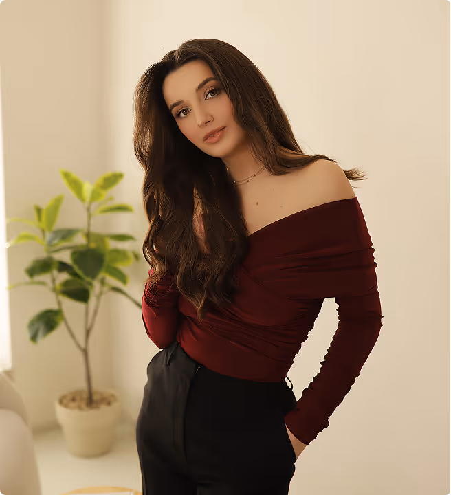Woman with long brown hair wearing a burgundy off-shoulder top and black pants, standing indoors near a potted plant.
