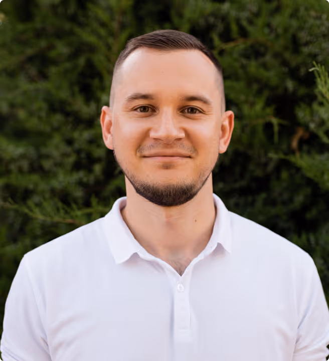 Smiling man with short hair and a neatly trimmed beard wearing a white polo shirt standing in front of green foliage.