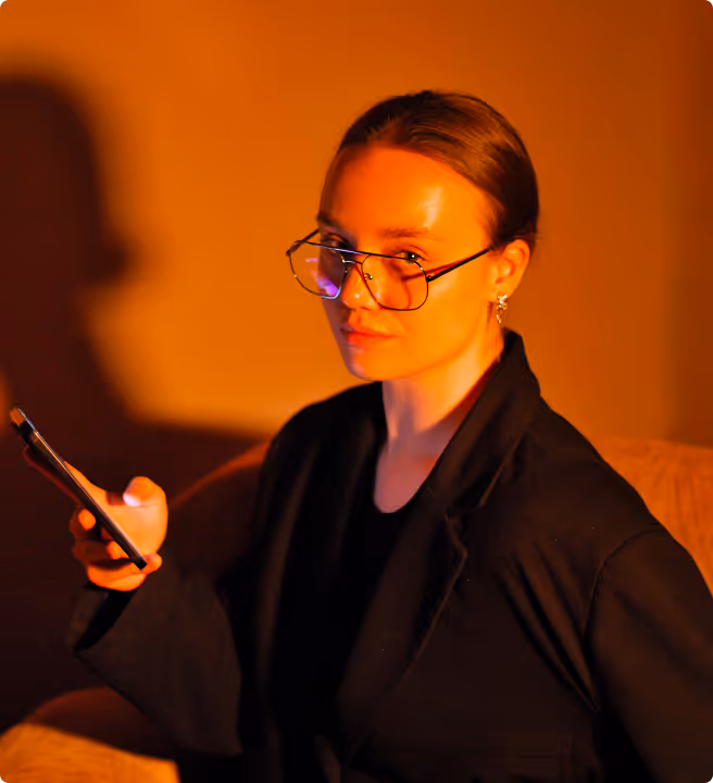 Woman with glasses wearing a black blazer holding a smartphone, illuminated by warm orange light.