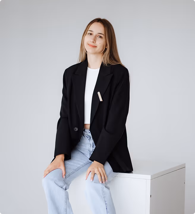 Young woman with long light brown hair wearing a black blazer, white top, and light blue jeans sitting on a white box against a plain gray background.