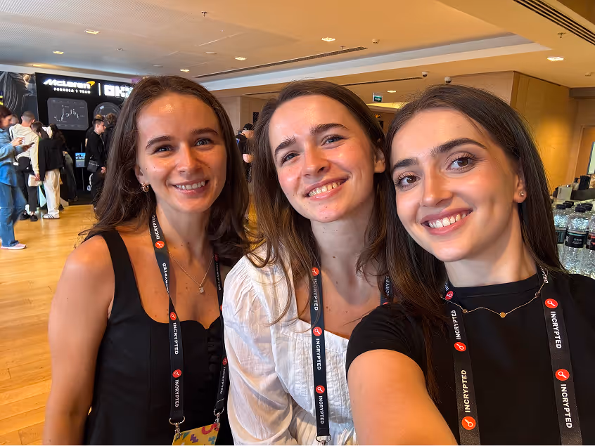 Three smiling young women with conference badges posing indoors in a well-lit room with people in the background.