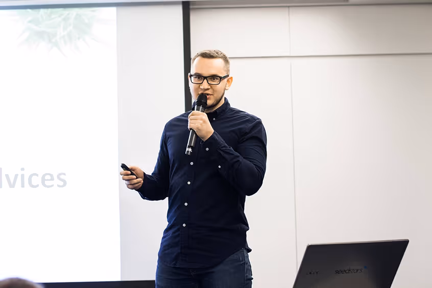 Man wearing glasses and a dark shirt speaking into a microphone during a presentation.