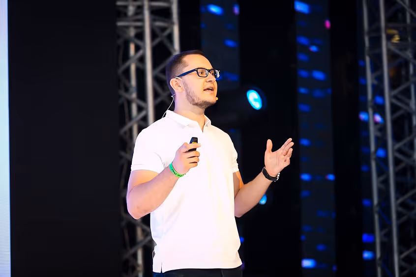 Man with glasses speaking on stage wearing a white polo shirt, holding a remote, with blue lights and metal truss structures in the background.