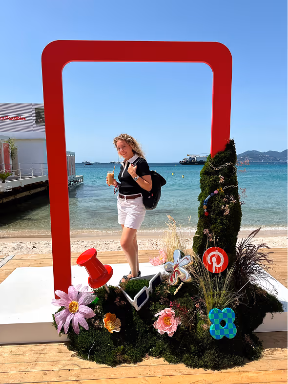Woman holding a drink standing inside a large red rectangular frame decorated with flowers and sunglasses, with a beach and ocean in the background.