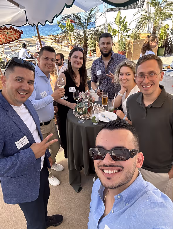 Group of seven young adults smiling and holding drinks around a table at an outdoor social gathering near the beach.