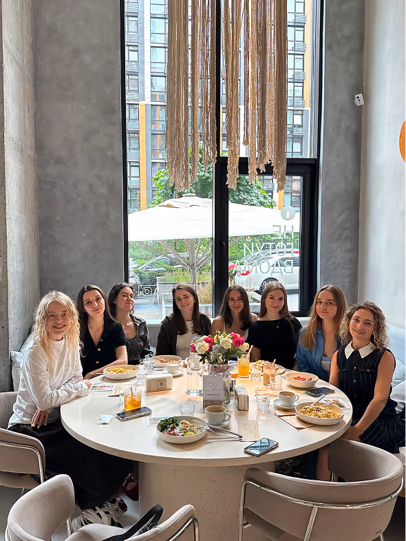 Group of seven women sitting around a round table with food and drinks in a modern café with large window and hanging decor.