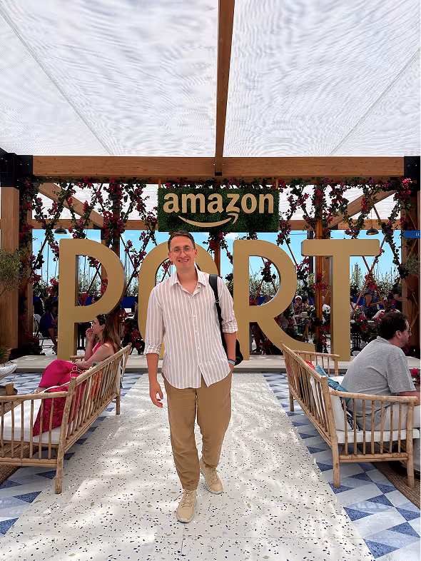 A man in glasses, striped shirt, and beige pants poses smiling in front of a large wood and floral decor sign reading 'amazon PORT' under a white canopy.