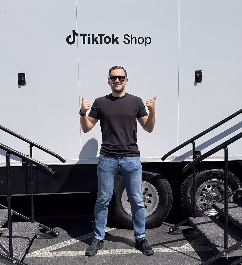 Man in black t-shirt, blue jeans, and sunglasses giving thumbs up standing between two staircases in front of a TikTok Shop trailer.