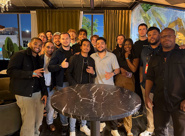 Group of diverse young adults smiling and posing together indoors around a marble-top table at night.