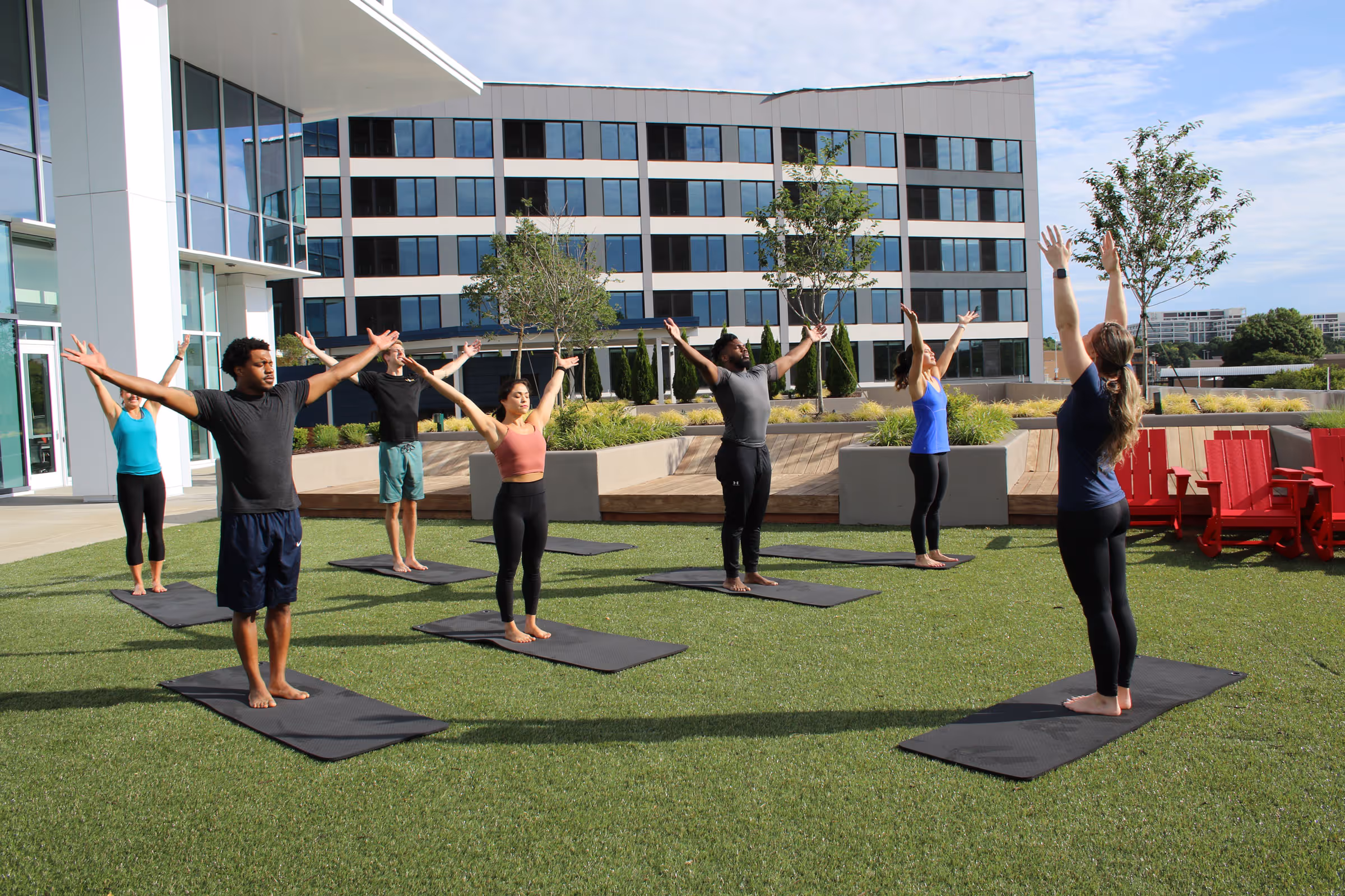 Group of six people practicing yoga outside on mats on a grass lawn, raising their arms upwards.