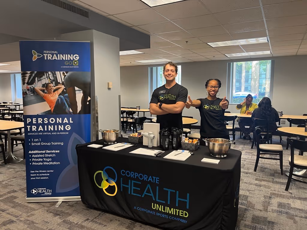 Two smiling fitness trainers standing behind a Corporate Health Unlimited table with promotional items and a personal training banner.