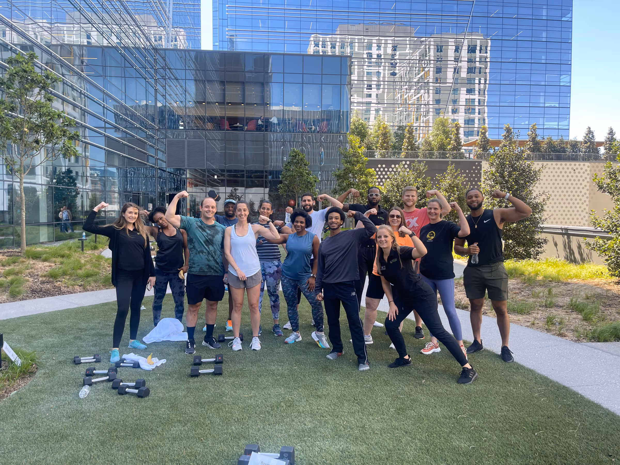 Group of people outdoors on grass in front of a modern glass building, flexing their arms after a workout with dumbbells on the ground.