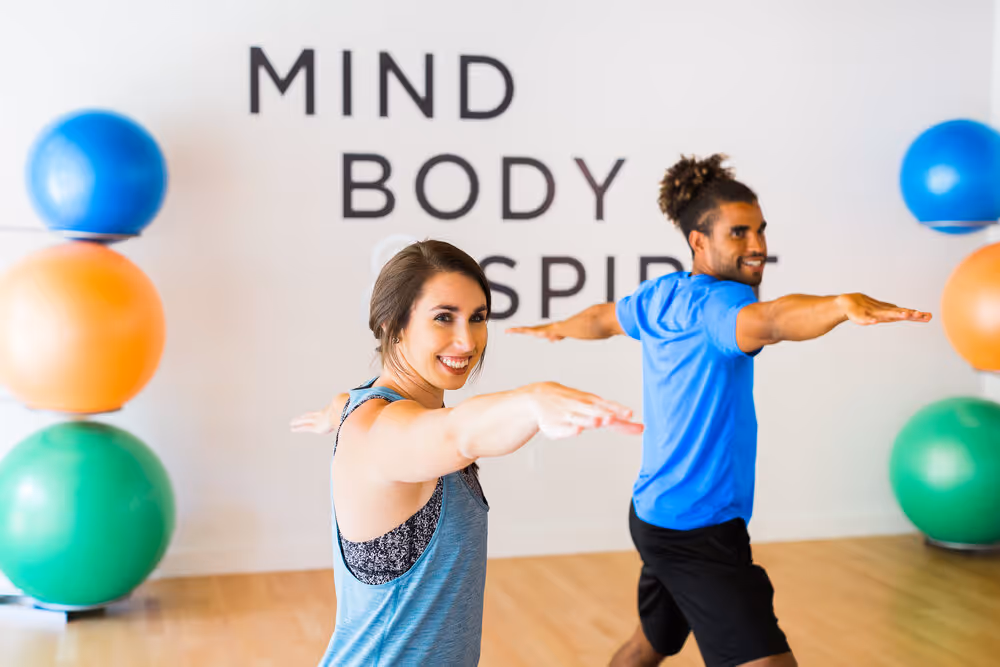 Two people practicing yoga with arms extended in a studio with exercise balls and text 'Mind Body Spirit' on the wall.