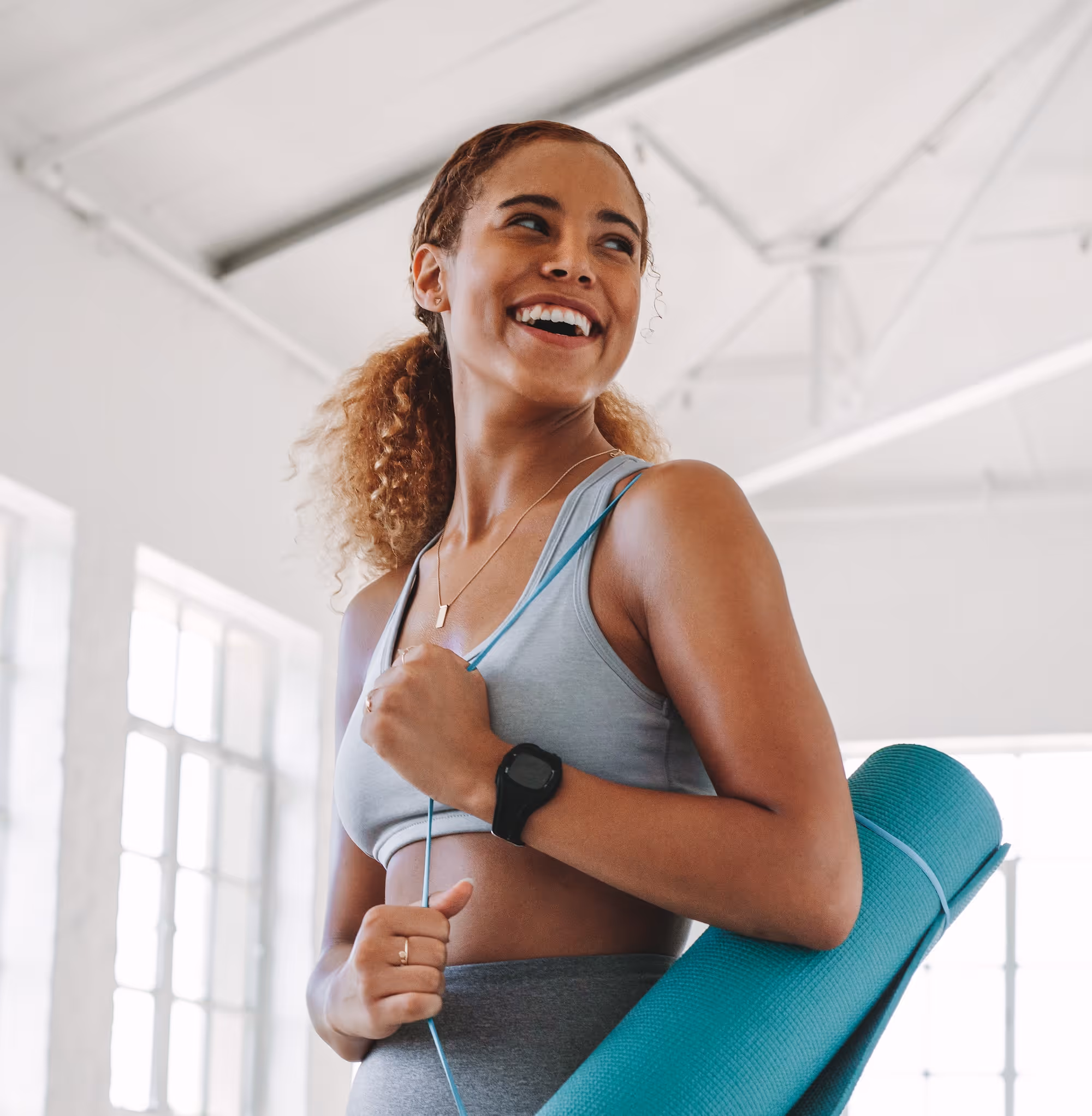 Smiling woman in athletic wear holding a rolled teal yoga mat over her shoulder in a bright studio.