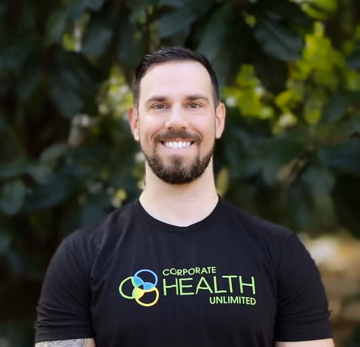 Smiling man with short dark hair and beard wearing a black t-shirt with Corporate Health Unlimited logo, standing outdoors with green foliage background.